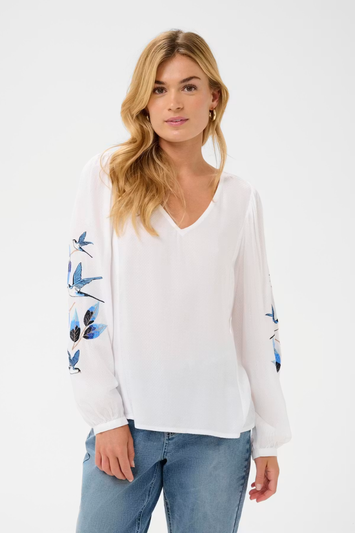 A woman wearing a white long-sleeve blouse with blue dragonfly embroidery on the sleeves, paired with blue jeans, against a plain white studio background.