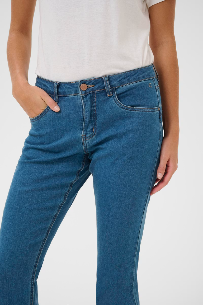 A close-up of a person wearing medium-wash blue denim jeans with a white t-shirt, showing the front pockets and waistband details against a plain light background.