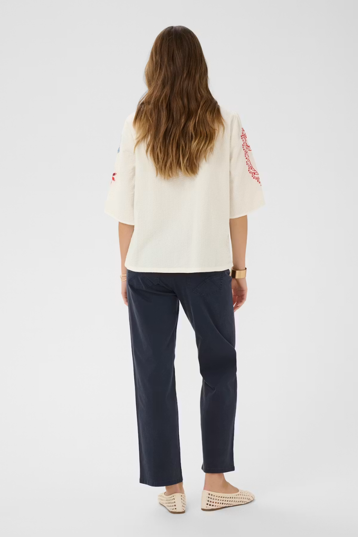 A woman is shown from the back wearing a cream-colored short-sleeve top with red embroidery on the sleeves, paired with dark navy trousers and beige woven flat shoes, against a plain light gray background.