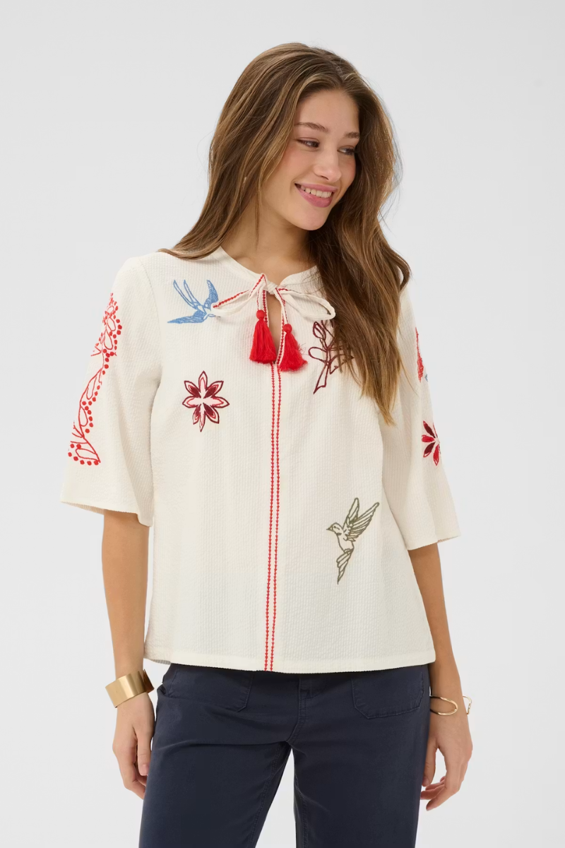 Woman wearing a white blouse with red floral embroidery and tassels on a plain background