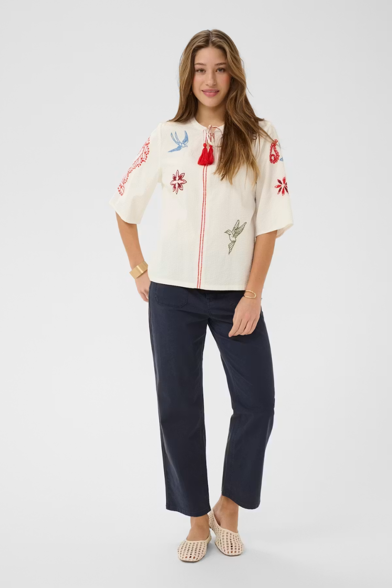 Woman wearing a white blouse with colorful embroidery and navy pants on a white background