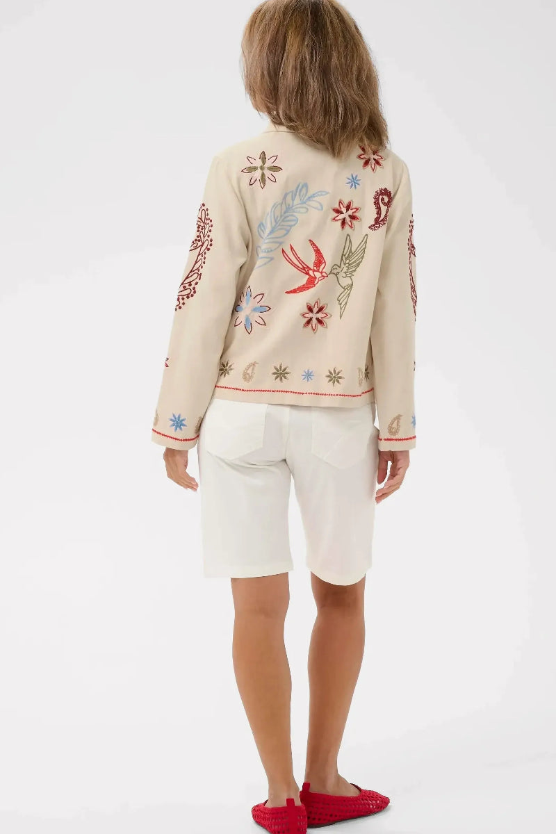 A child model shown from the back wearing a cream-colored embroidered cardigan with colorful bird and floral motifs, white shorts, and red slip-on shoes, against a light gray studio background.