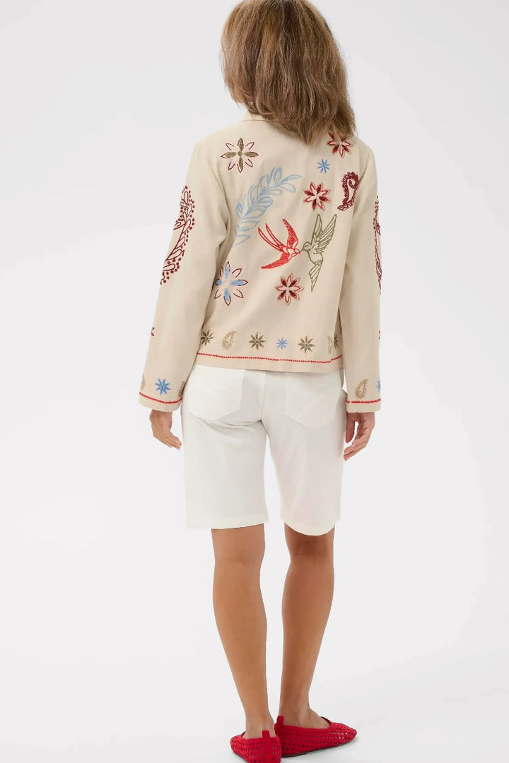 A child model shown from the back wearing a cream-colored embroidered cardigan with colorful bird and floral motifs, white shorts, and red slip-on shoes, against a light gray studio background.