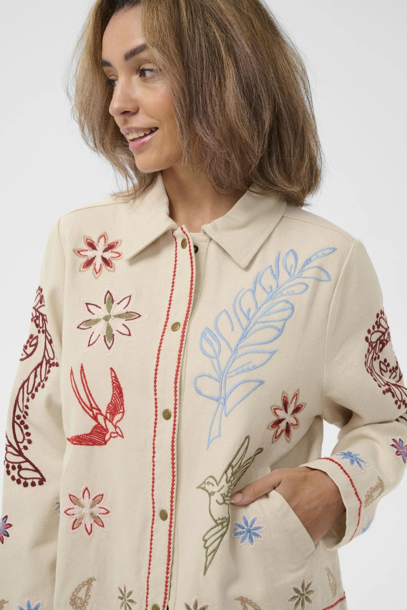 A woman wearing a cream-colored embroidered button-up shirt jacket with colorful floral and bird motifs, photographed against a plain light gray studio background.