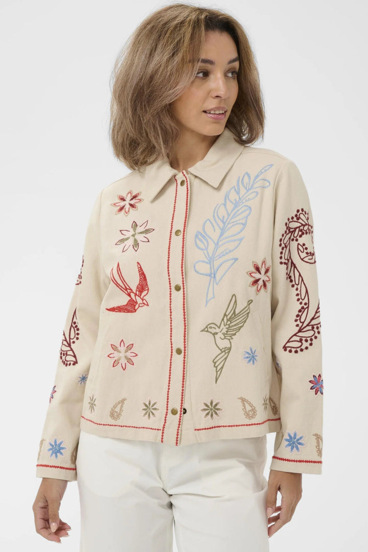 A woman wearing a cream-colored embroidered jacket with red, blue, and green floral and bird motifs, paired with white pants, against a plain light gray background.