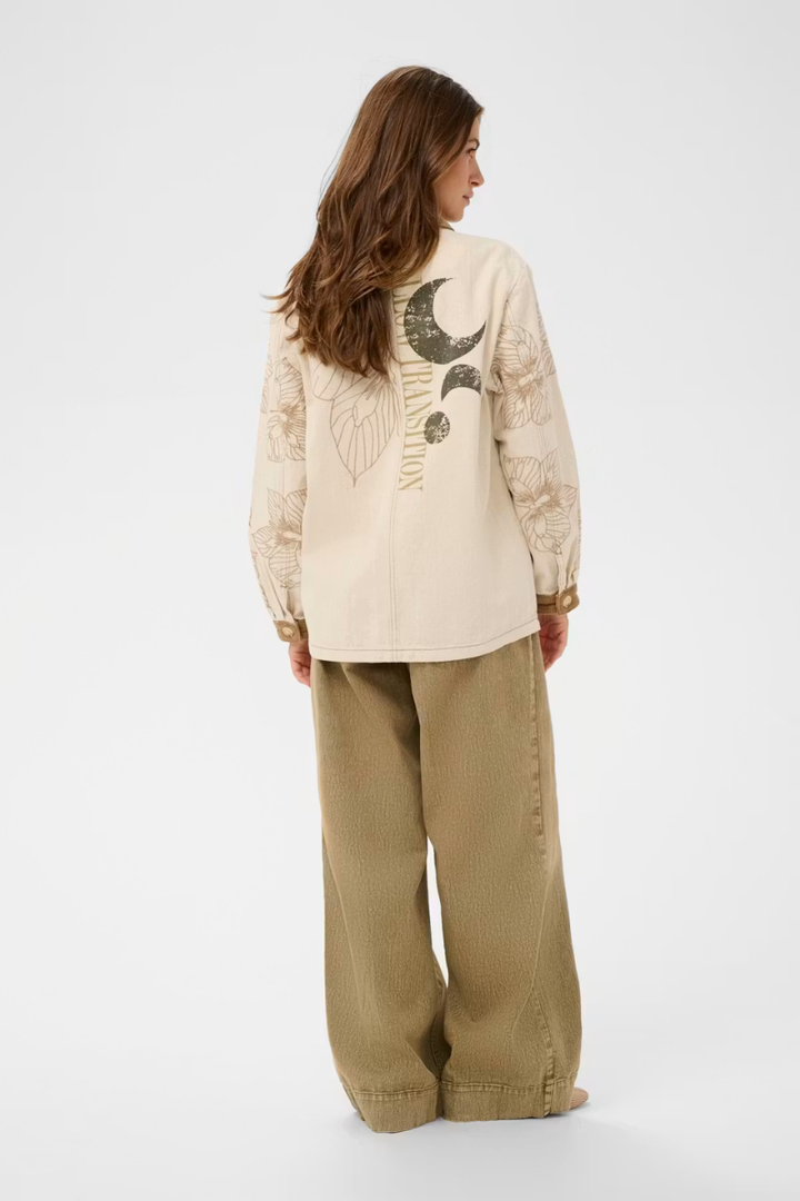 A woman shown from the back wearing a cream-colored long-sleeve shirt with a black moon and leaf graphic on the back, paired with wide-leg beige pants, against a plain white studio background.