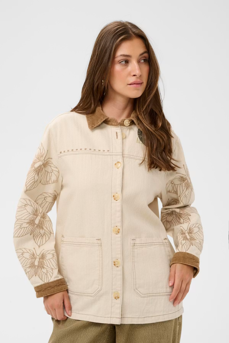 A woman wearing a cream-colored button-up jacket with brown floral embroidery on the sleeves, brown corduroy collar and cuffs, and gold-tone buttons, set against a plain light gray background.