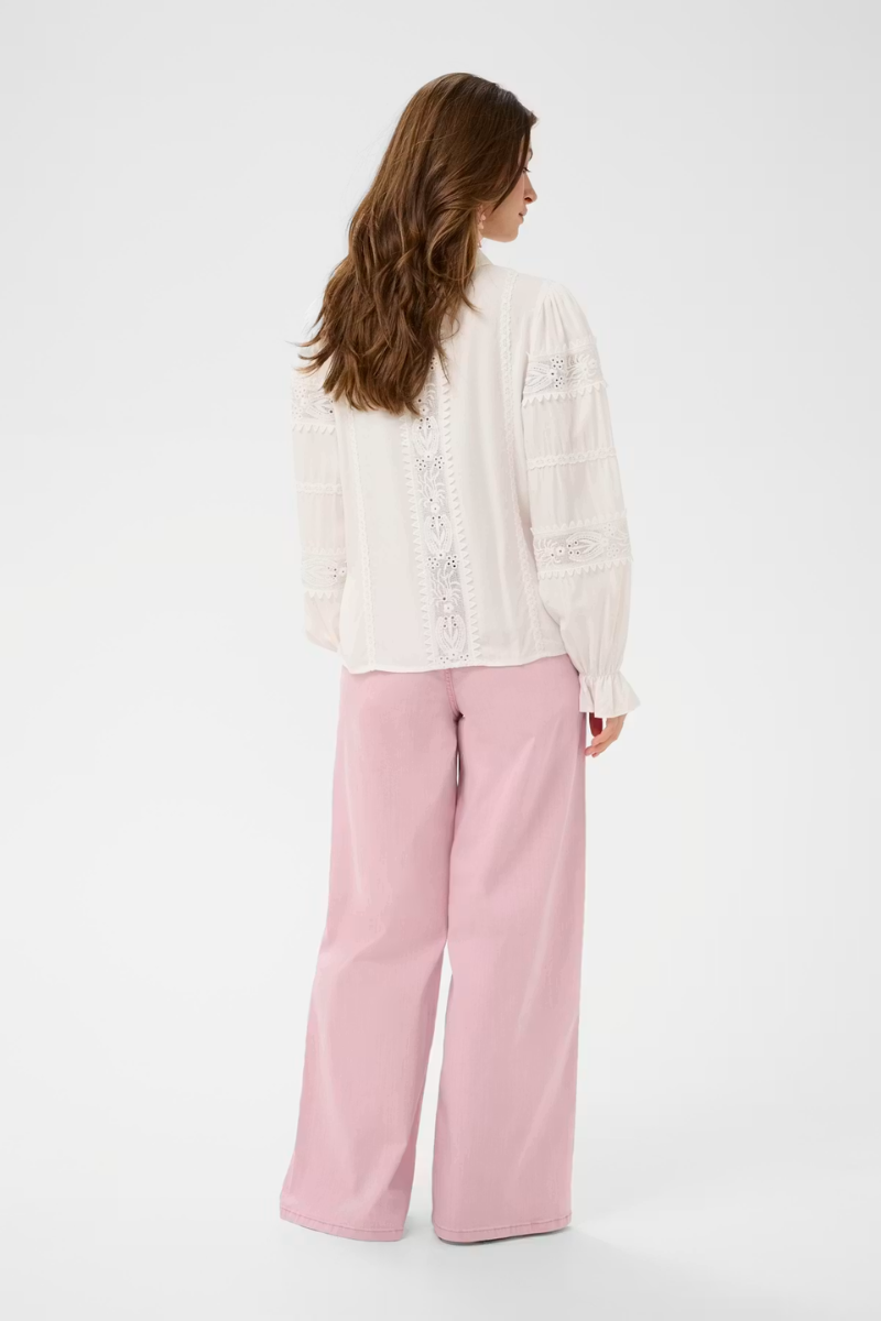 A woman shown from the back wearing a white long-sleeve blouse with lace detailing and light pink wide-leg trousers, photographed against a plain white studio background.