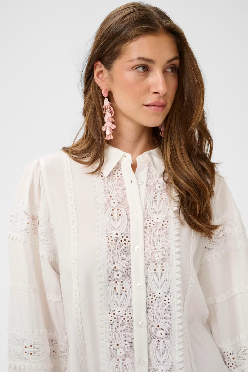 A woman wearing a white long-sleeve button-up blouse with intricate floral eyelet embroidery down the front, paired with pink dangling earrings, against a plain light gray background.