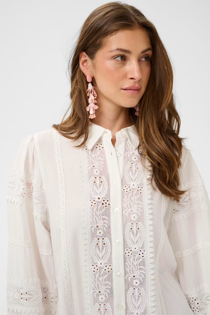 A woman wearing a white long-sleeve button-up blouse with intricate floral eyelet embroidery down the front, paired with pink dangling earrings, against a plain light gray background.