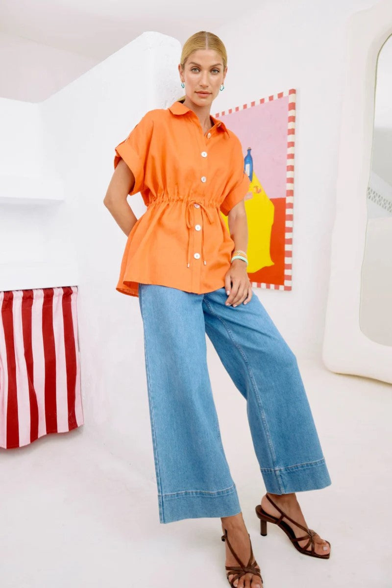 Woman wearing an orange shirt and blue jeans standing in a room with colorful abstract art on the wall.