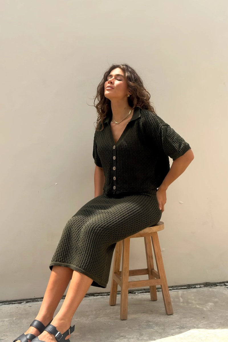 Woman sitting on a wooden stool wearing a black and green patterned outfit against a plain wall.