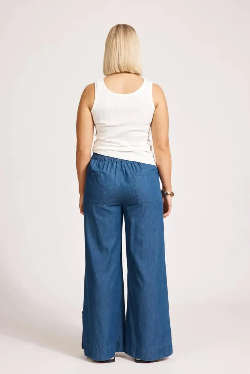 A woman viewed from the back wearing a white sleeveless top and wide-leg blue denim pants against a plain light background.
