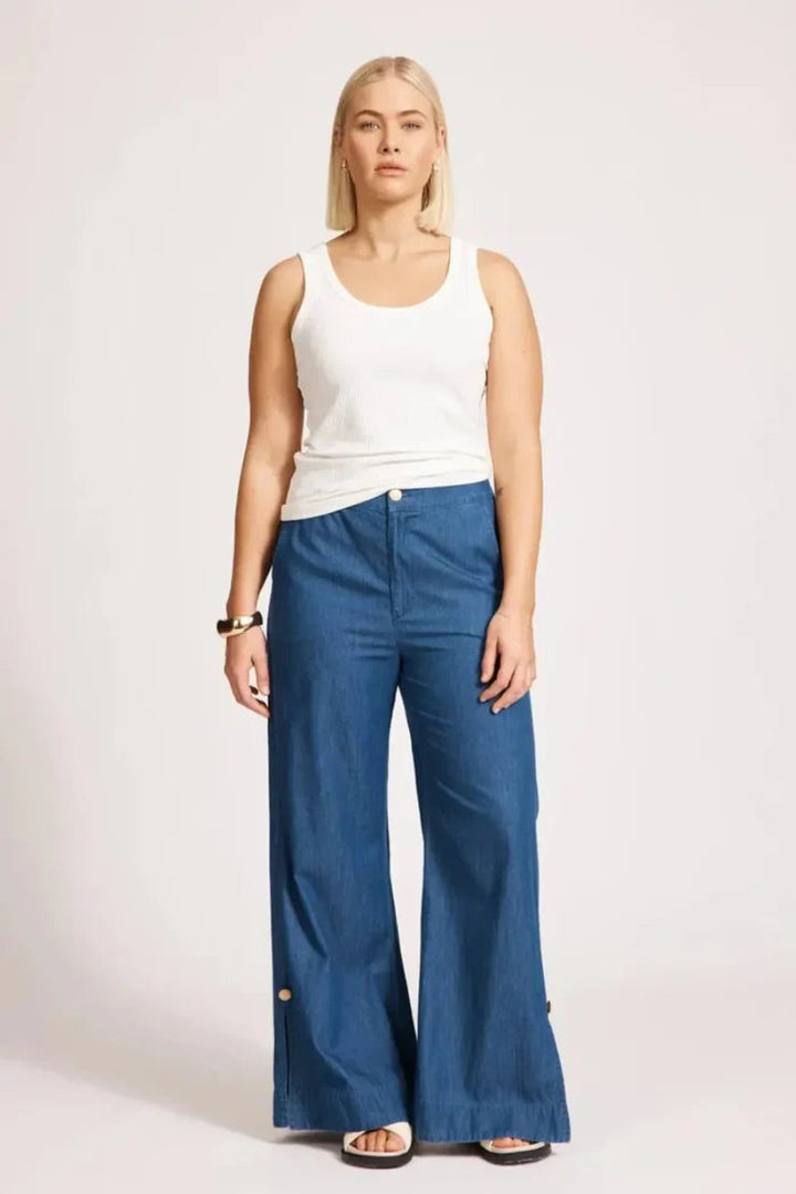 A woman modeling a pair of wide-leg blue jeans with a white ribbed tank top, standing against a light gray studio background.