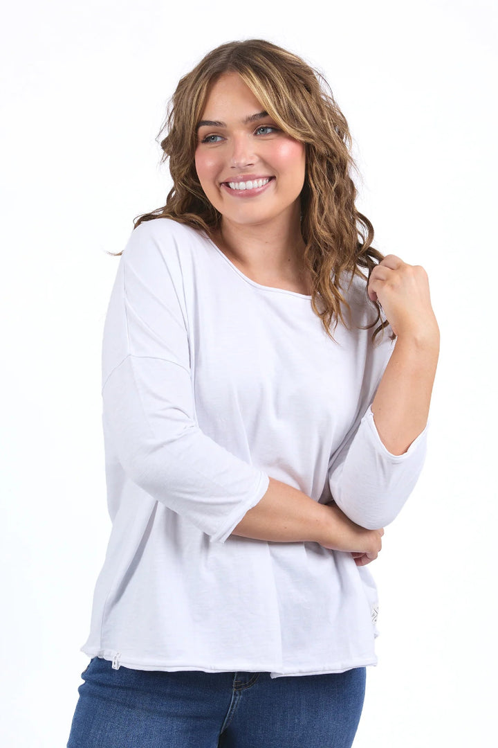 A smiling woman with wavy brown hair wearing a white three-quarter sleeve top and blue jeans, posing against a solid white background.
