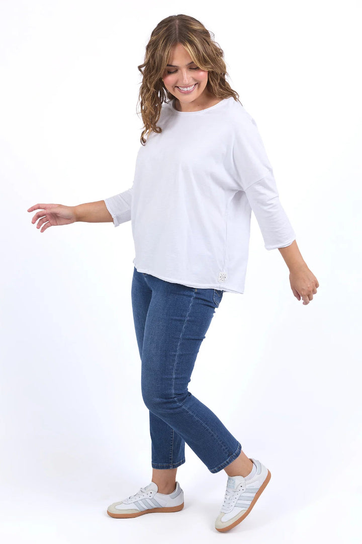 A woman modeling a white three-quarter sleeve top, blue skinny jeans, and white sneakers against a plain white background.