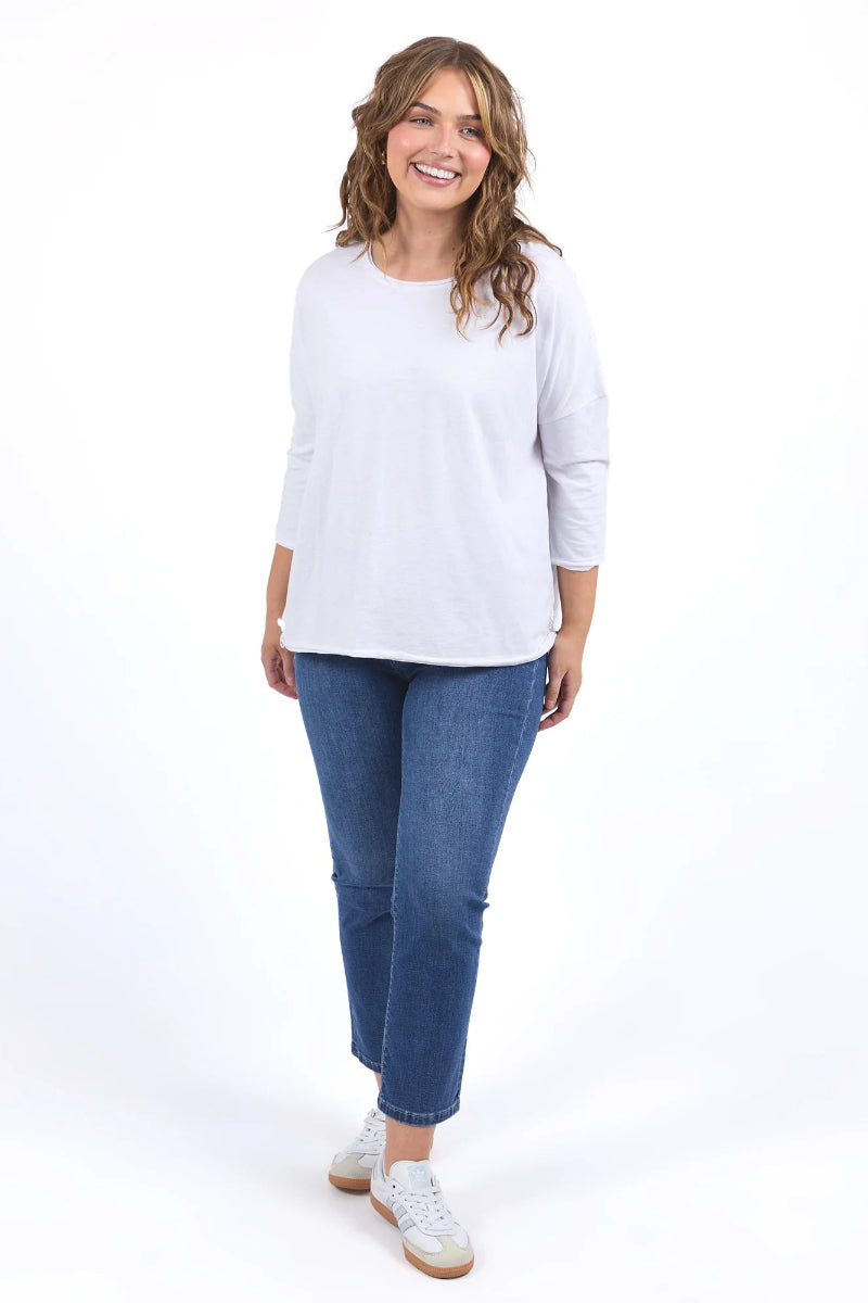 A woman modeling a white three-quarter sleeve top, blue cropped jeans, and white sneakers against a plain white studio background.
