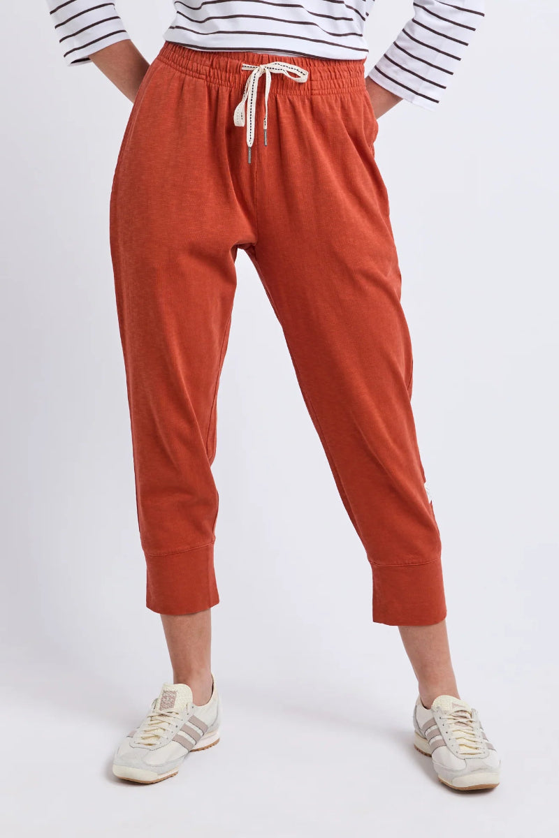 A person wearing rust-orange cropped jogger pants with an elastic drawstring waist, paired with a striped long-sleeve top and white sneakers, shown against a light gray studio background.
