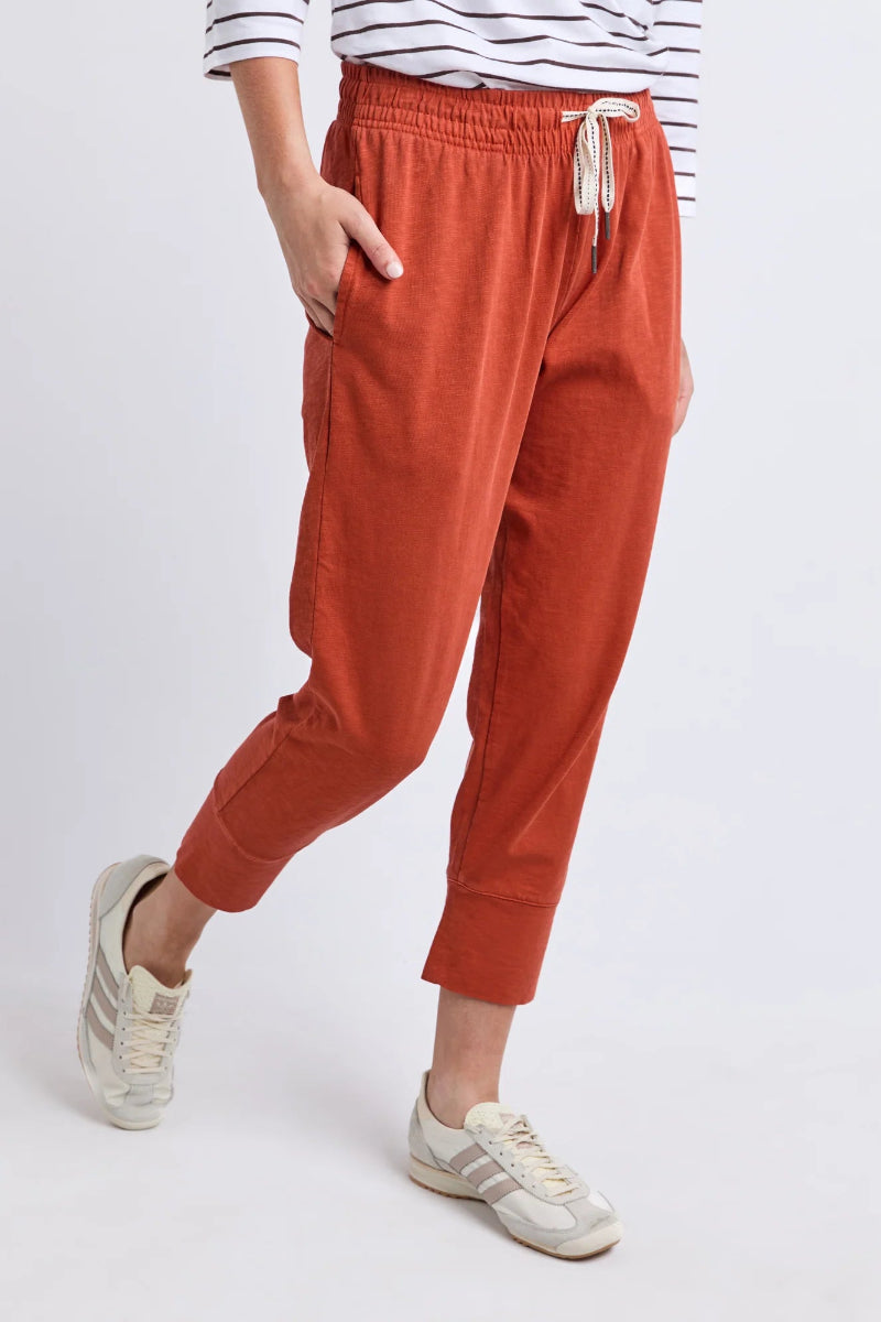 A person wearing rust-orange drawstring jogger pants with side pockets, styled with a striped top and white sneakers, shown from the waist down against a light gray studio background.
