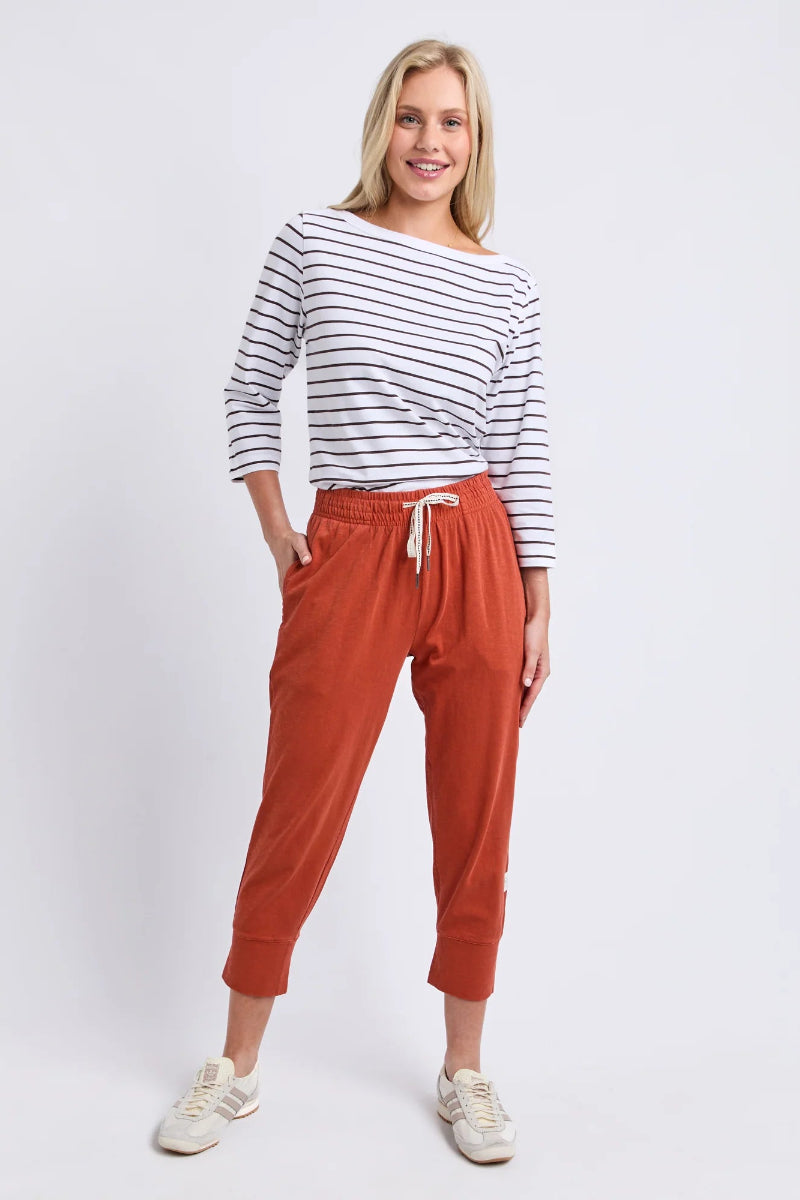 A woman modeling a casual outfit consisting of a white long-sleeve top with thin navy horizontal stripes and rust-orange cropped jogger pants with a white drawstring, paired with white sneakers, against a light gray studio background.