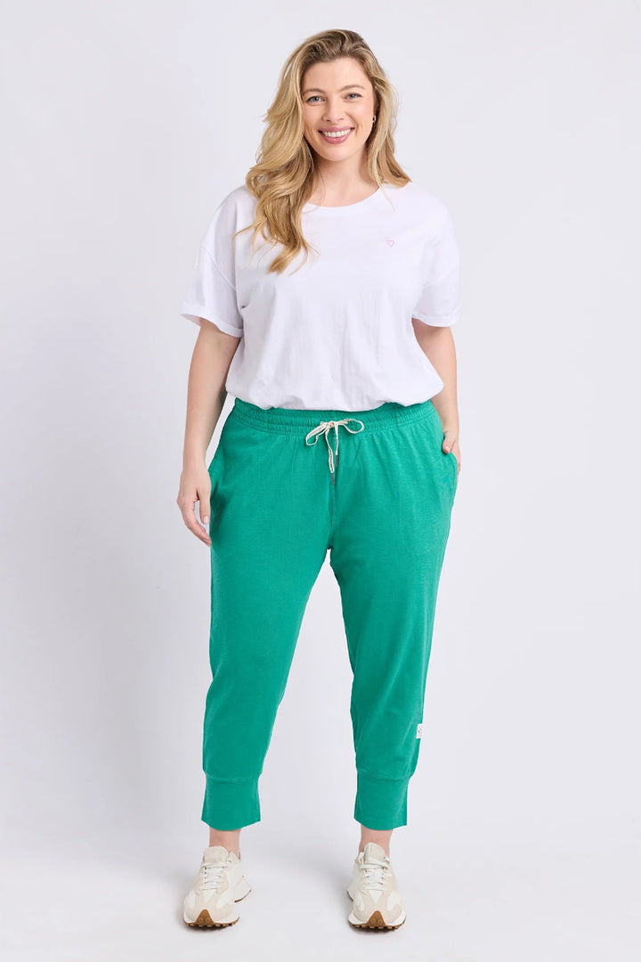 A woman modeling bright green jogger pants with a white drawstring, styled with a white t-shirt and white sneakers against a light gray studio background.