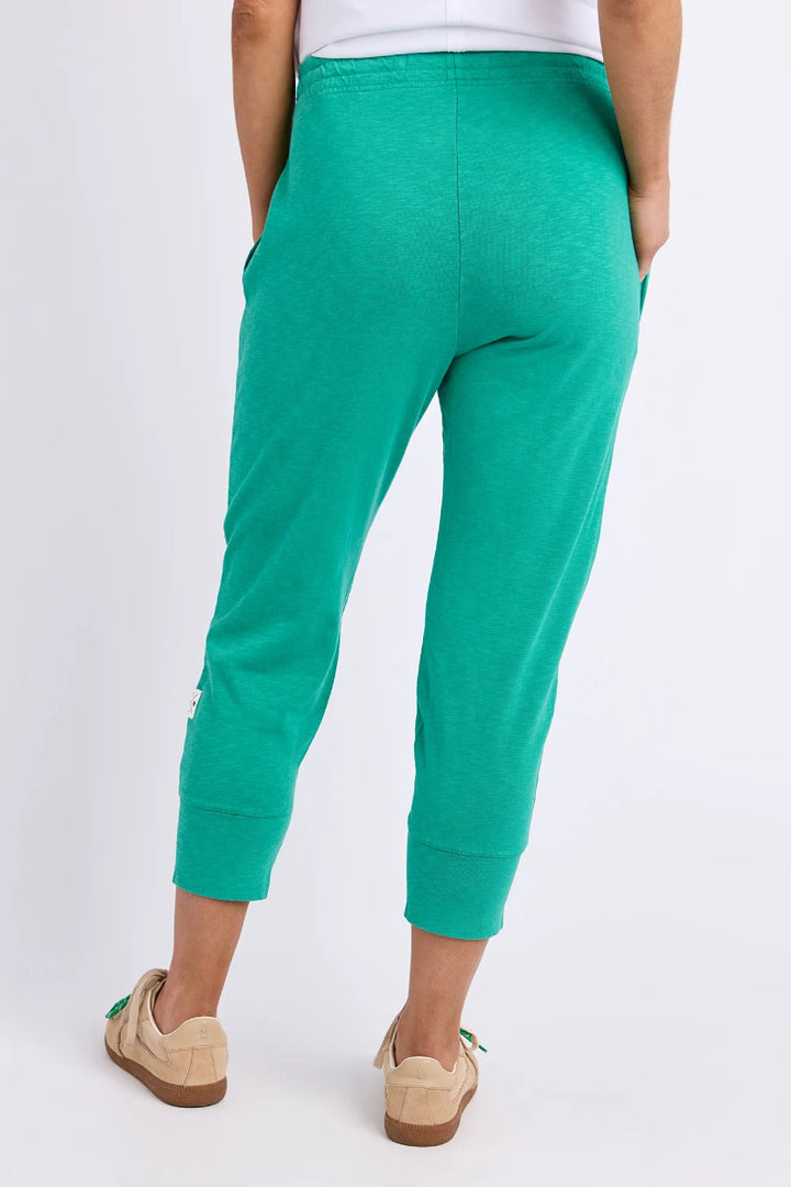 A person shown from the back wearing bright green cropped sweatpants with side pockets, a white top, and beige slip-on shoes against a plain light gray studio background.