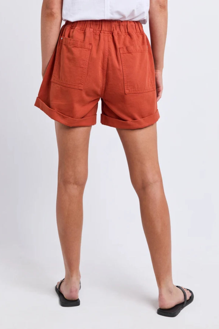 A person shown from the back wearing high-waisted orange-brown shorts with a gathered elastic waistband, two large patch pockets, and rolled cuffs, paired with a white top and black sandals on a light gray studio background.