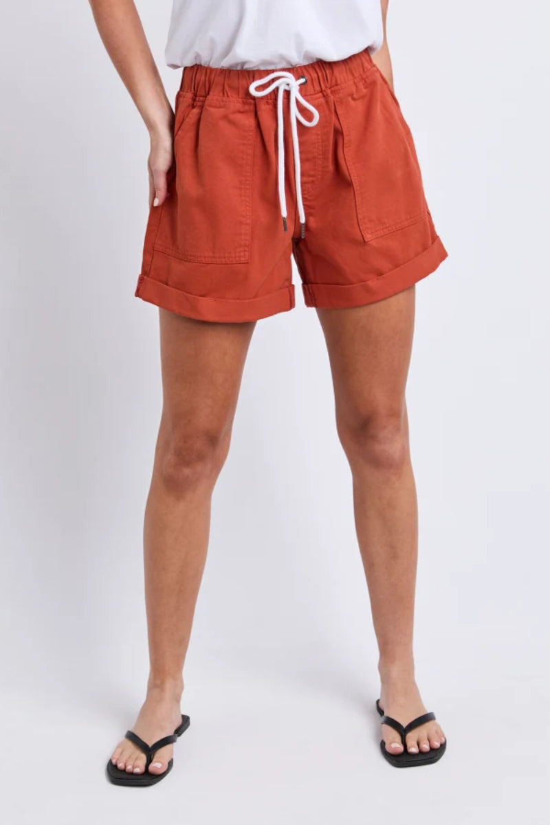 A person wearing rust-orange drawstring shorts with a white top and black flip-flops, standing against a light gray studio background.