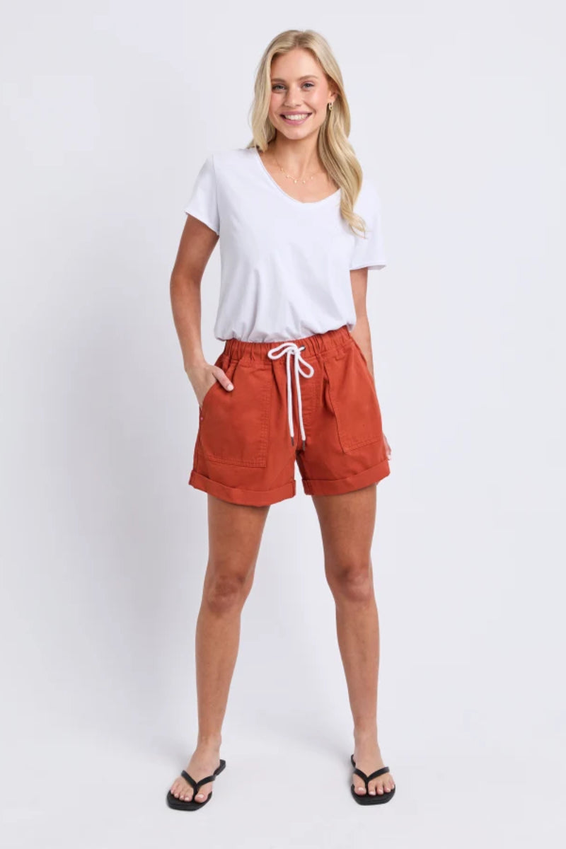 A woman modeling a white short-sleeve V-neck t-shirt and rust-orange drawstring shorts against a light gray studio background.