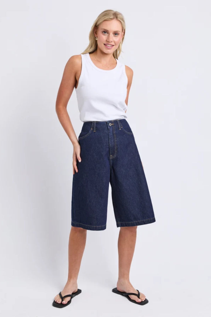 A woman modeling dark wash wide-leg denim shorts with a white sleeveless tank top and black flip-flops, standing against a light gray studio background.