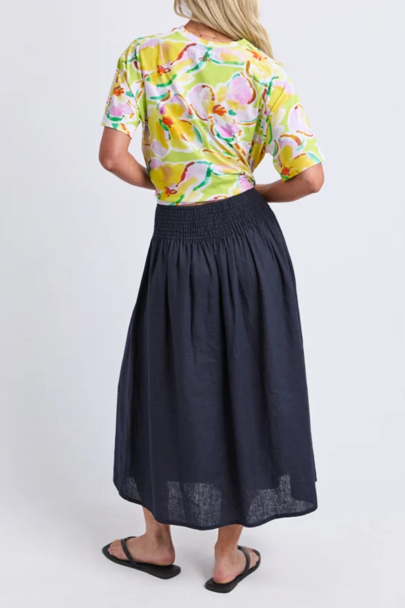 A woman shown from the back wearing a bright yellow floral short-sleeve top and a dark navy midi skirt, paired with black sandals, against a light gray studio background.