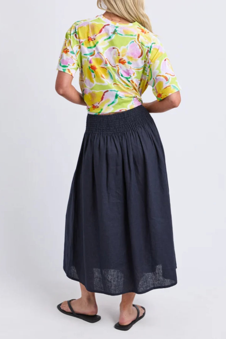 A woman shown from the back wearing a bright yellow floral short-sleeve top and a dark navy midi skirt, paired with black sandals, against a light gray studio background.