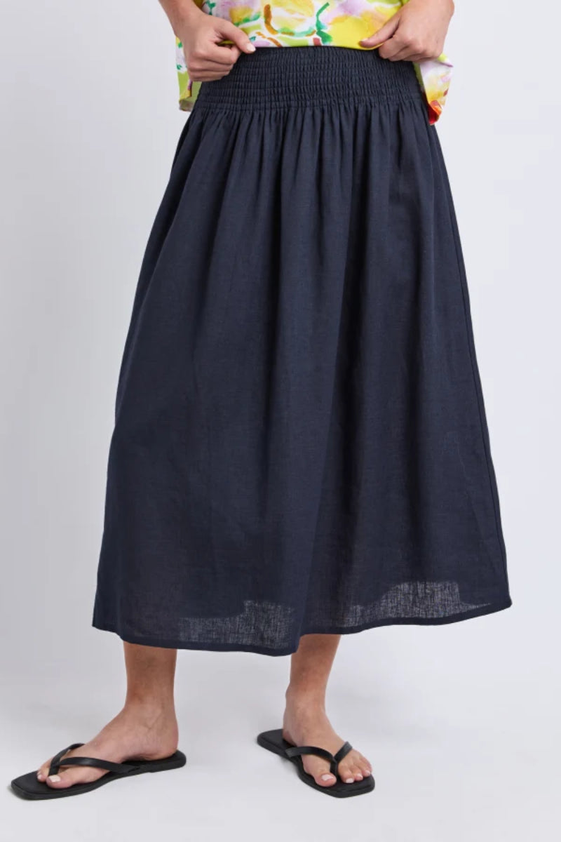 A person wearing a dark navy midi skirt with a smocked waistband, paired with a colorful floral top and black flip-flop sandals, shown against a light gray studio background.