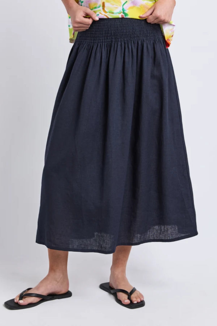 A person wearing a dark navy midi skirt with a smocked waistband, paired with a colorful floral top and black flip-flop sandals, shown against a light gray studio background.