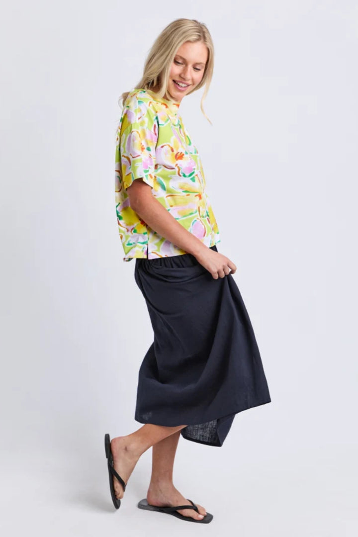 A woman modeling a short-sleeve button-up shirt with a colorful flamingo print on a white background, paired with a dark navy midi skirt and black flip-flops, against a light gray studio backdrop.
