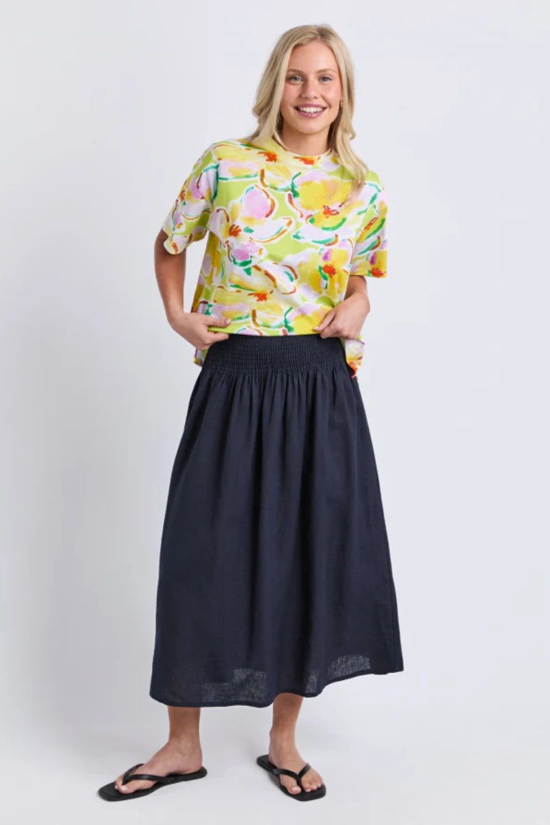 A woman modeling a short-sleeve yellow and green floral print top paired with a dark navy midi skirt and black flip-flop sandals, standing against a light gray studio background.