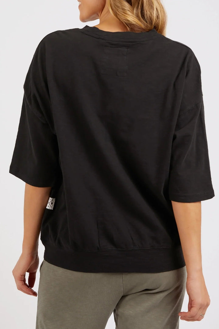 A woman shown from the back wearing a black oversized short-sleeve crewneck sweatshirt and olive green pants against a light gray studio background.