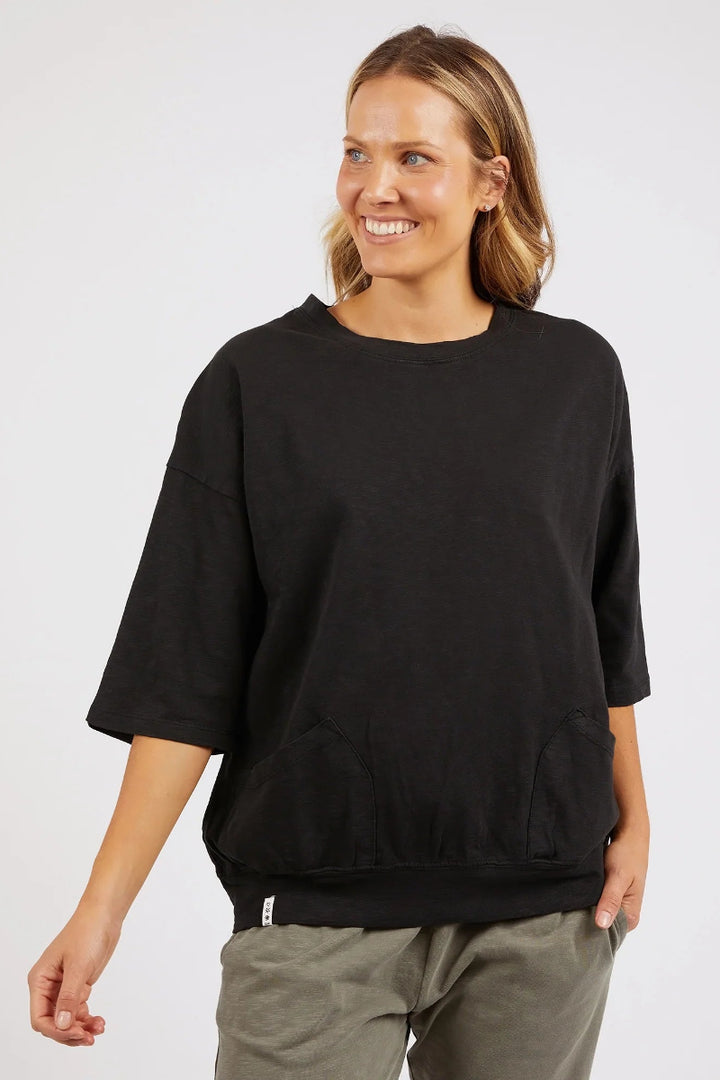 A woman wearing an oversized black crewneck sweatshirt with three-quarter sleeves and front pockets, paired with olive green pants, photographed against a light gray studio background.
