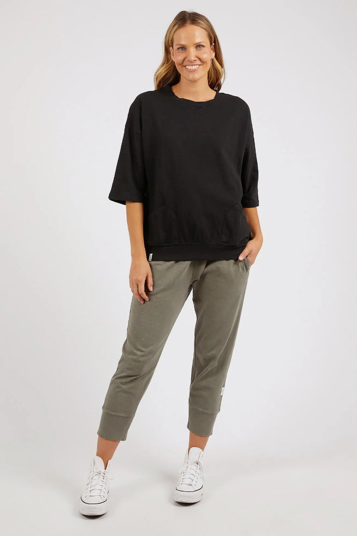 A woman modeling a black short-sleeve crewneck sweatshirt and olive green cropped pants with white sneakers, standing against a light gray studio background.