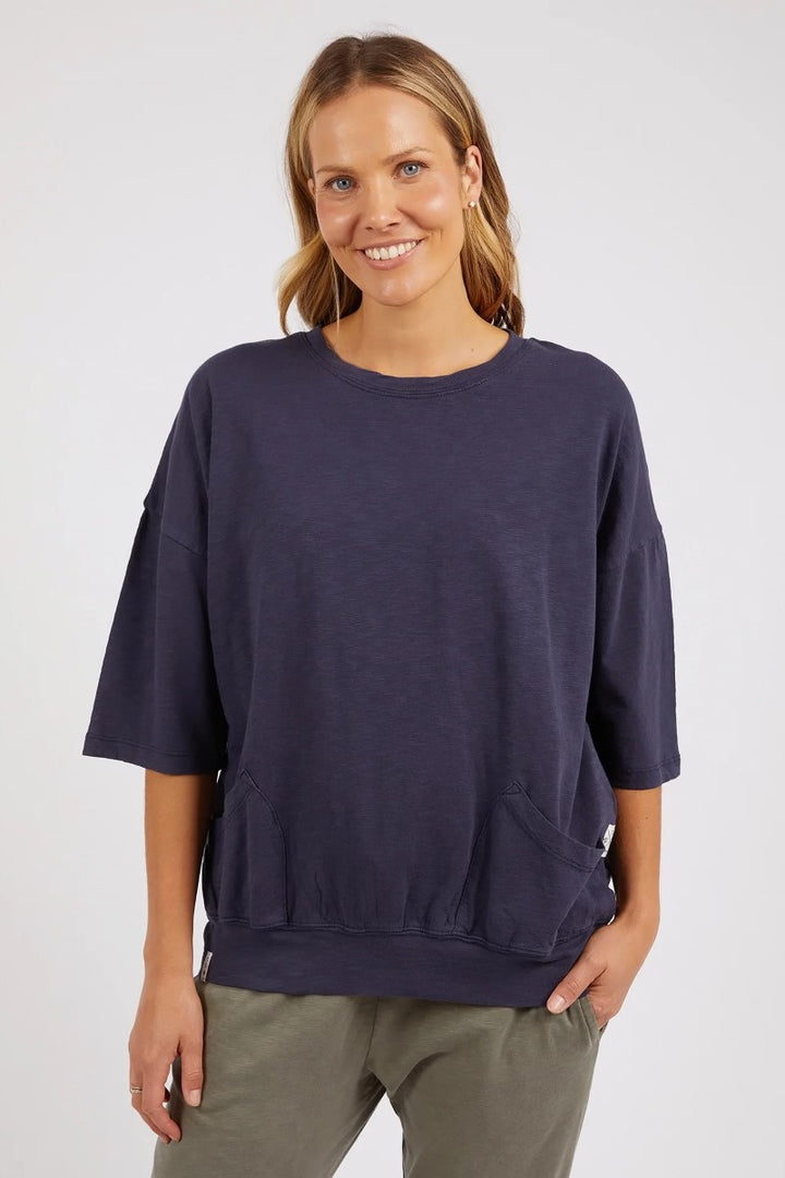 A woman smiling, wearing a navy-colored, 3/4 sleeve Elm Mazie Sweat with front pockets and a high rounded neckline.