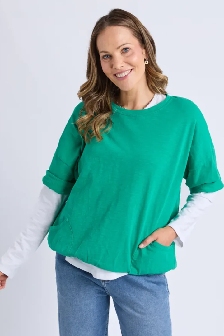 Woman wearing a green sweater over a white shirt with blue jeans on a light gray background