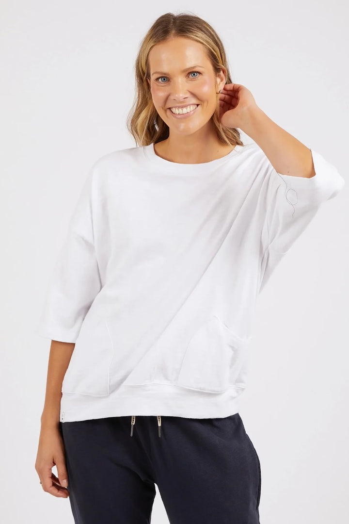 A woman wearing a white oversized sweatshirt with 3/4 length sleeves and front pockets, paired with dark pants.