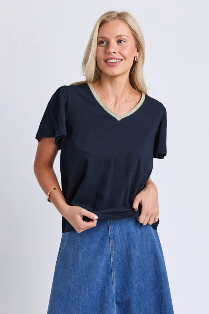 A woman modeling a navy blue V-neck short-sleeve top with light green trim, paired with a blue denim skirt, against a light gray studio background.