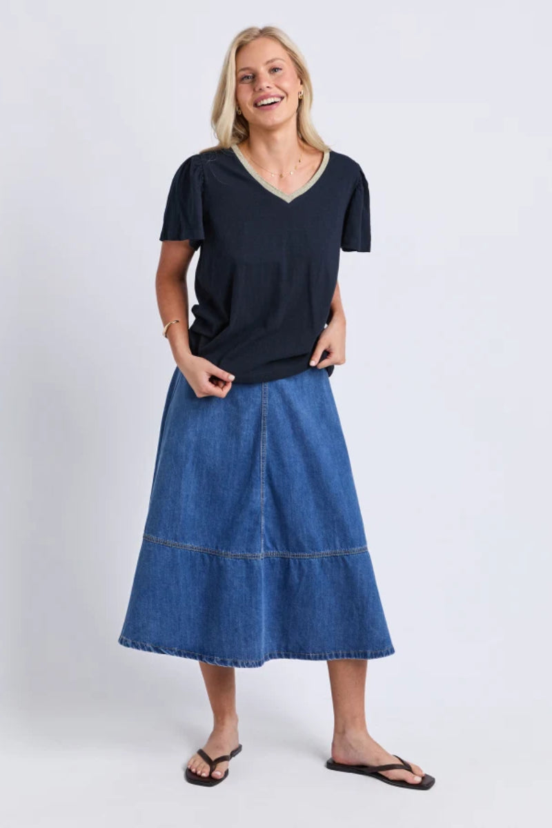 A woman modeling a dark navy blue short-sleeve V-neck top and a blue denim midi skirt, paired with black flip-flops, against a light gray studio background.