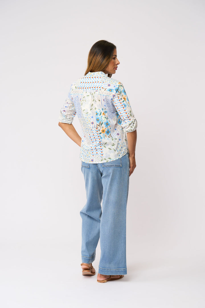 A woman is shown from the back wearing a white embroidered blouse with colorful floral and geometric patterns, paired with light blue wide-leg jeans and brown sandals, against a plain light gray studio background.
