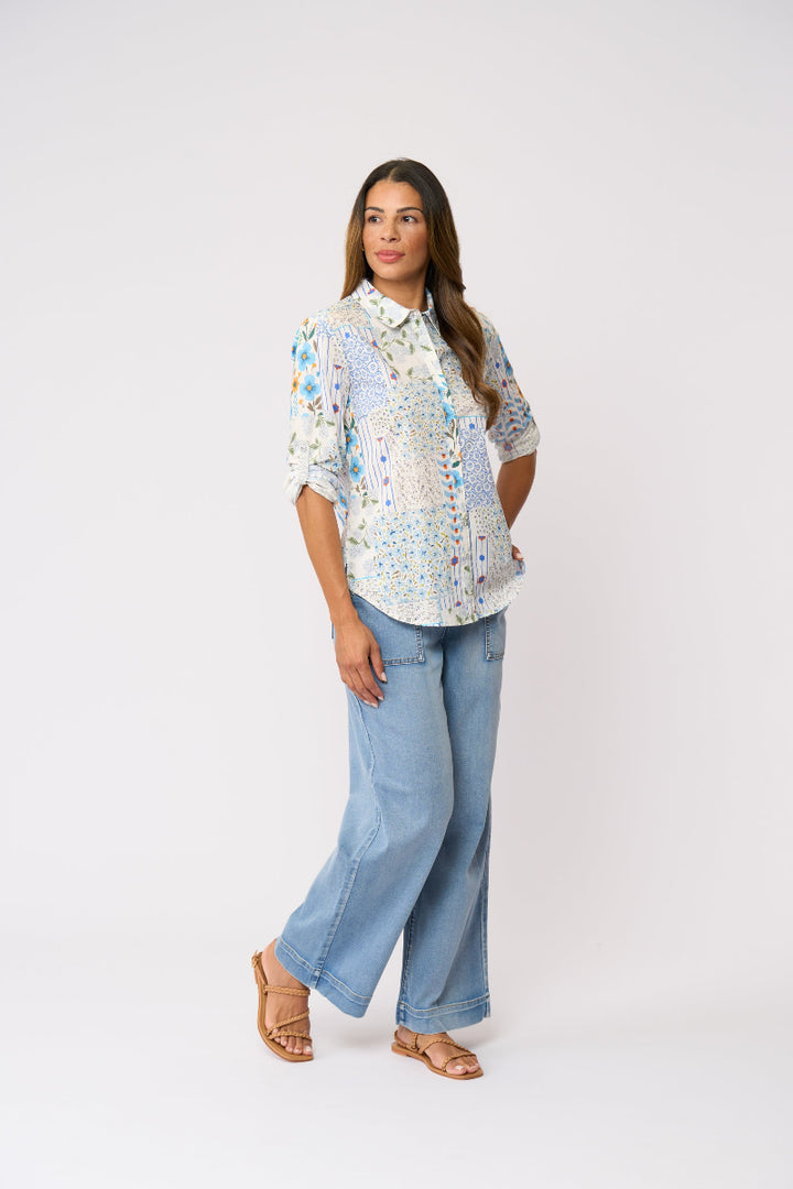 A woman modeling a white button-up shirt with a colorful blue and yellow abstract print, paired with light-wash wide-leg jeans and tan sandals, against a plain white studio background.