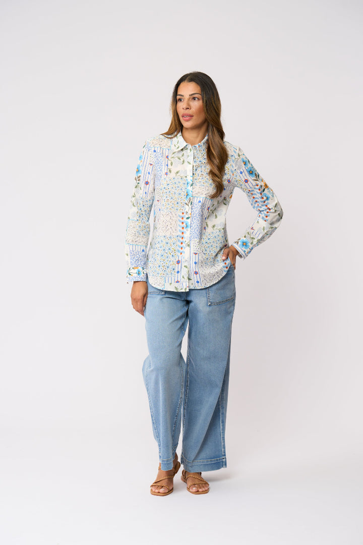 A woman modeling a long-sleeve button-up shirt with a dense multicolor floral and abstract print on a white base, paired with light blue wide-leg jeans and brown sandals, against a plain white studio background.