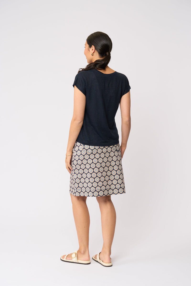 A woman is shown from the back wearing a navy blue short-sleeve t-shirt and a beige skirt with a black geometric hexagon pattern, paired with beige slide sandals, against a plain white studio background.