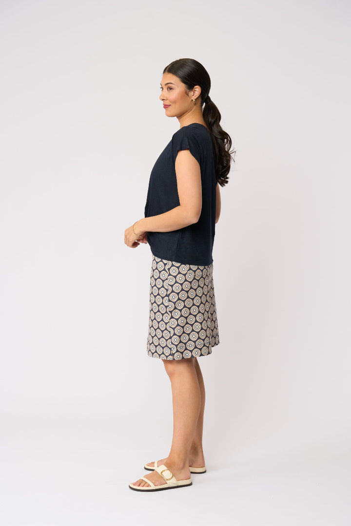 A woman shown in profile wearing a dark navy short-sleeve top and a beige skirt with a dark geometric pattern, standing against a plain light gray studio background.