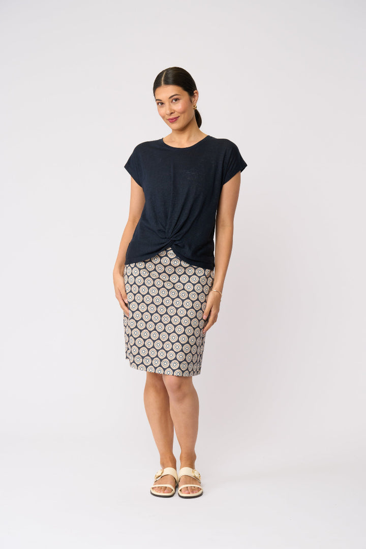 A woman modeling a short-sleeve dark navy top with a twisted front detail and a knee-length skirt featuring a beige and black geometric pattern, paired with beige slide sandals, against a light gray studio background.